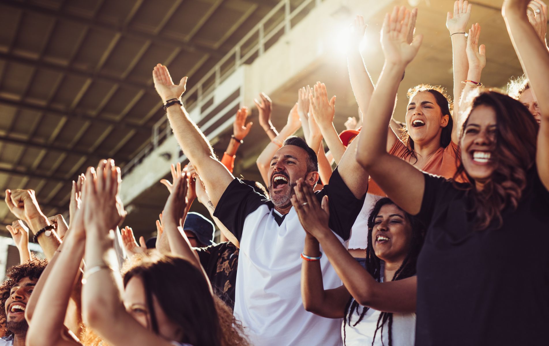 Crowd of diverse sports fans in a stadium cheering with hands raised and smiles on their faces in bright light.