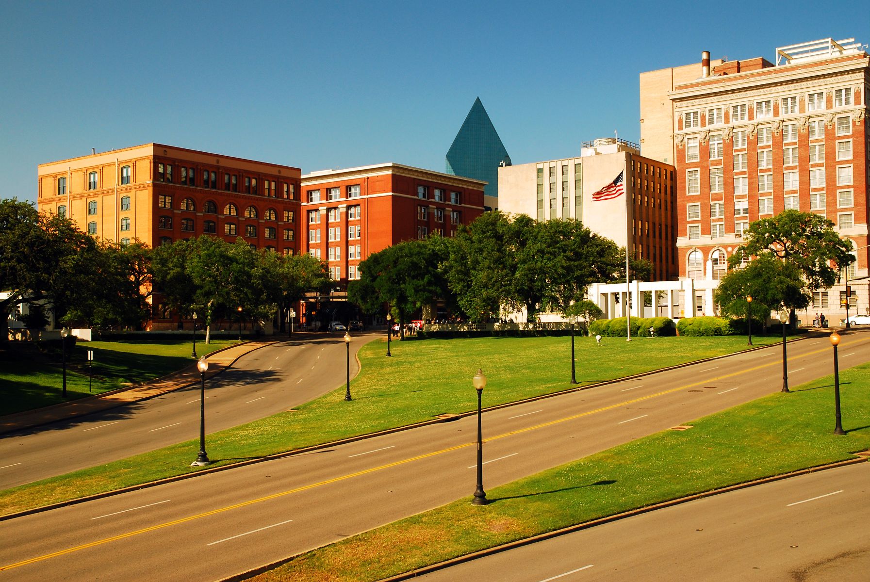 City street lined with red brick buildings, green trees, and a curved roadway under a clear blue sky.