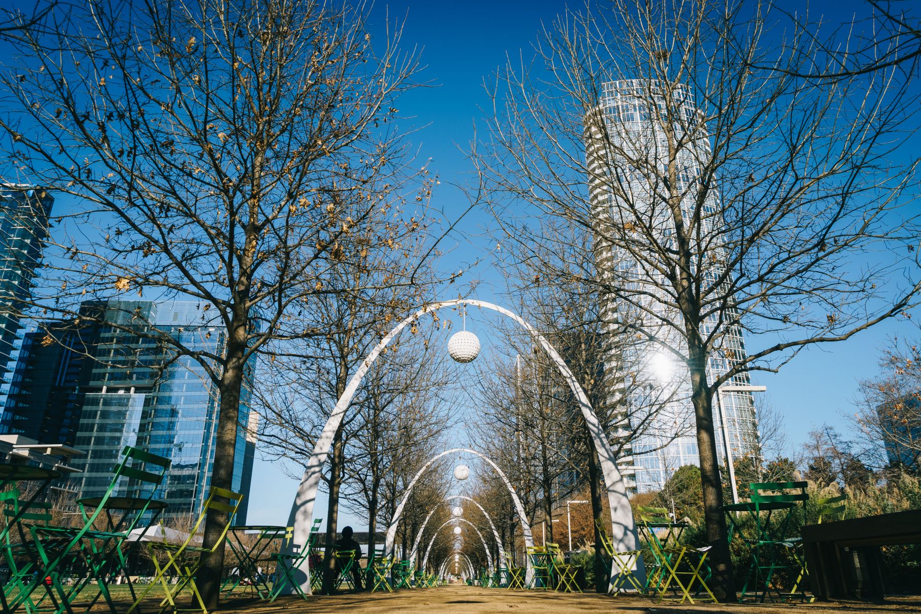 Row of white arches with hanging spheres along a tree-lined path in a city park, with modern glass skyscrapers in the background.